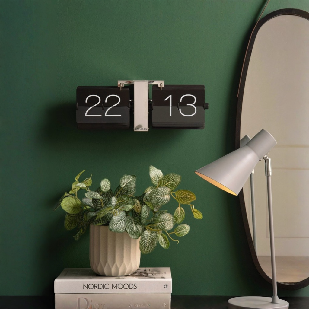 Flip retro airport style clock on a green background styled next to a mirror and with coffee table books, vase with green plants and table lamp 