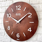 Brown wall clock with white numbers and 'EQS' branding on a white brick wall background