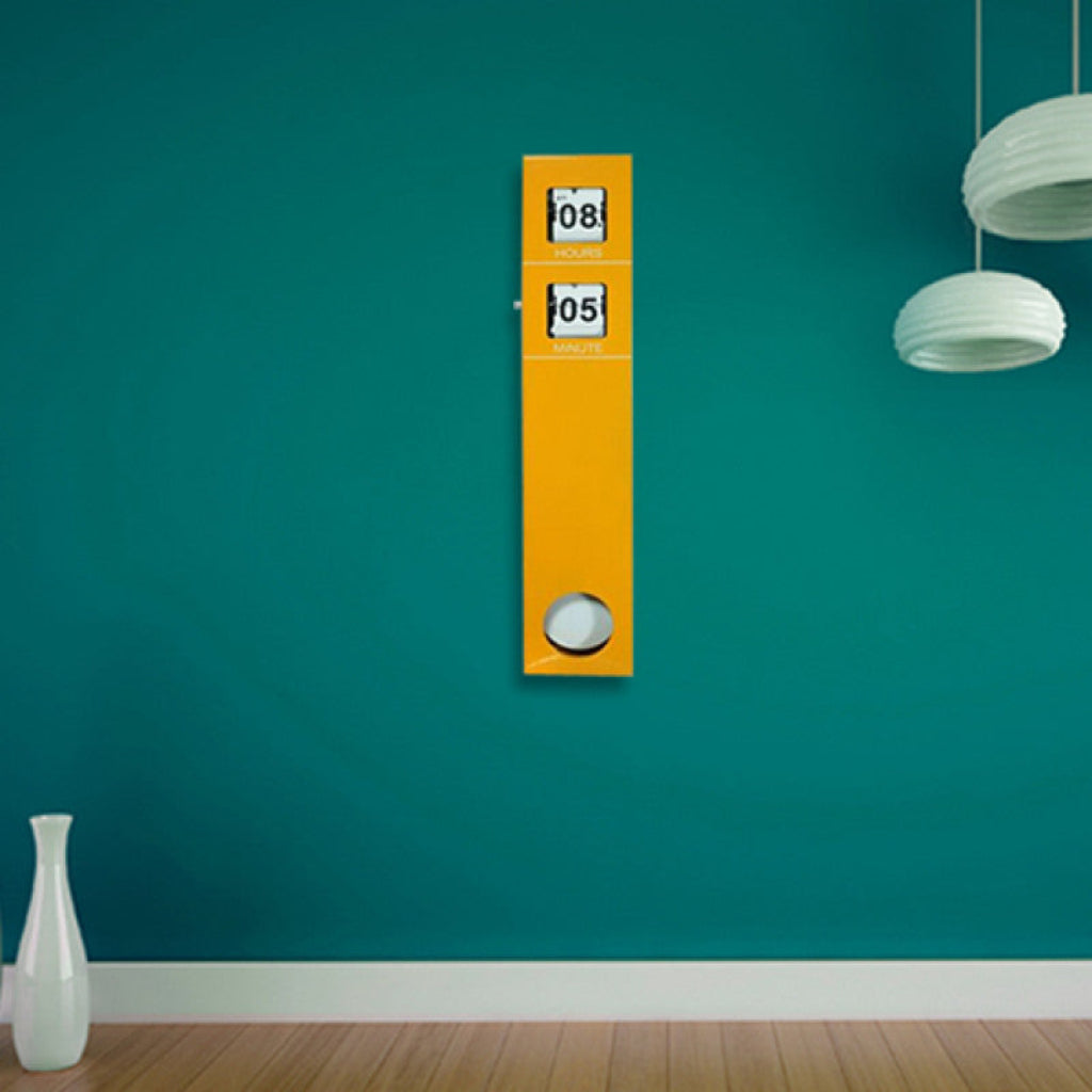 Yellow vertical clock on a teal wall with a white vase and lamp in the foreground.