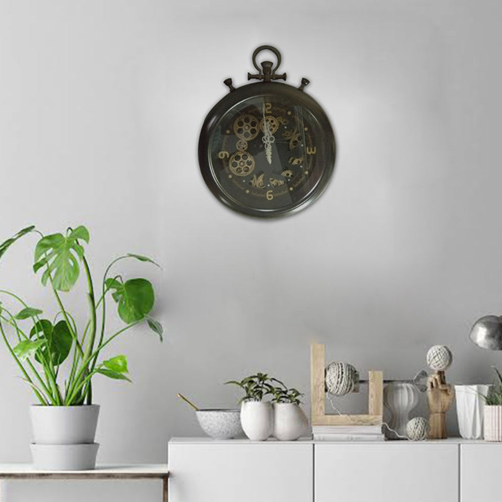 Decorative wall clock with mechanical design on a gray wall, next to a plant and small decorative items.