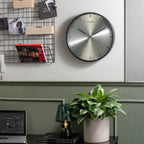 Modern wall clock on a wall with a plant and desk setup below.