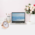 Laptop on a desk with a clock, pen holder, and flowers against a light pink wall.
