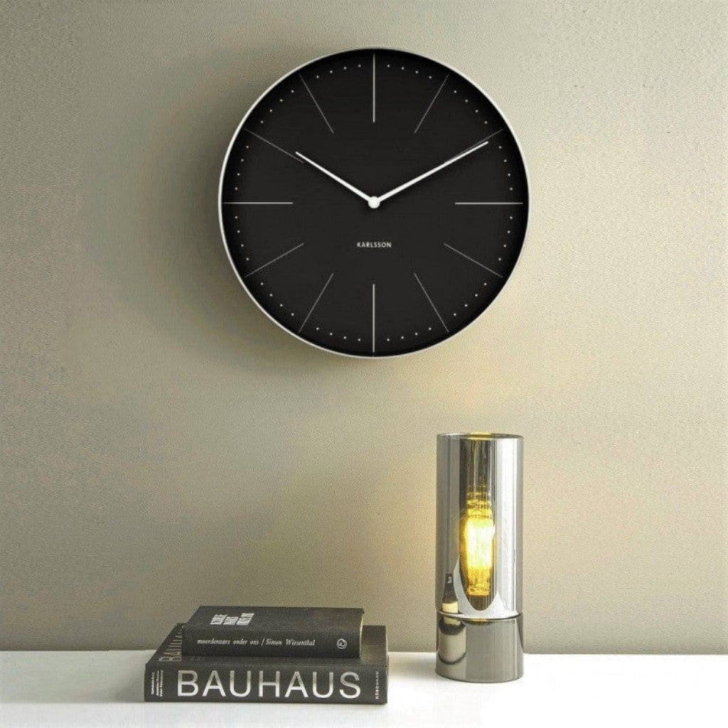 Black wall clock on a light gray wall with a small lamp and books below.