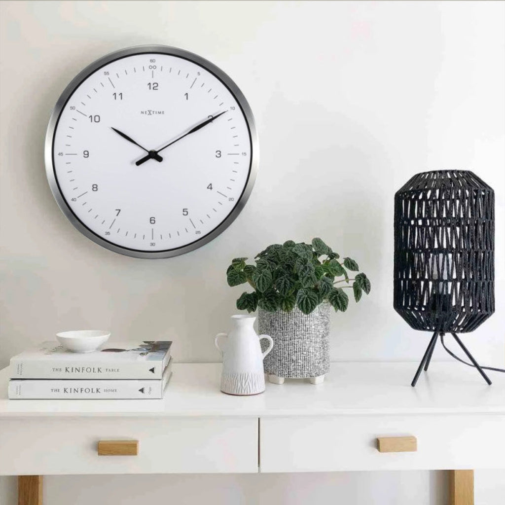 Wall clock on a white wall above a shelf with books, a plant, and a lamp.