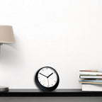 Black clock on a shelf with books and a lamp in the background