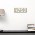 Digital clock displaying the time on a white wall with a lamp and books below.