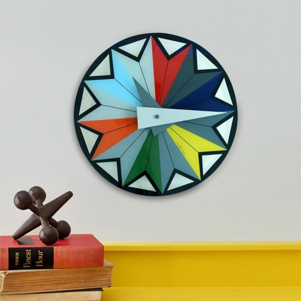 Colorful geometric wall clock on a white wall with books and a sculpture below.
