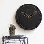 Black wall clock with rose gold hands on a white wall, next to a small potted plant.