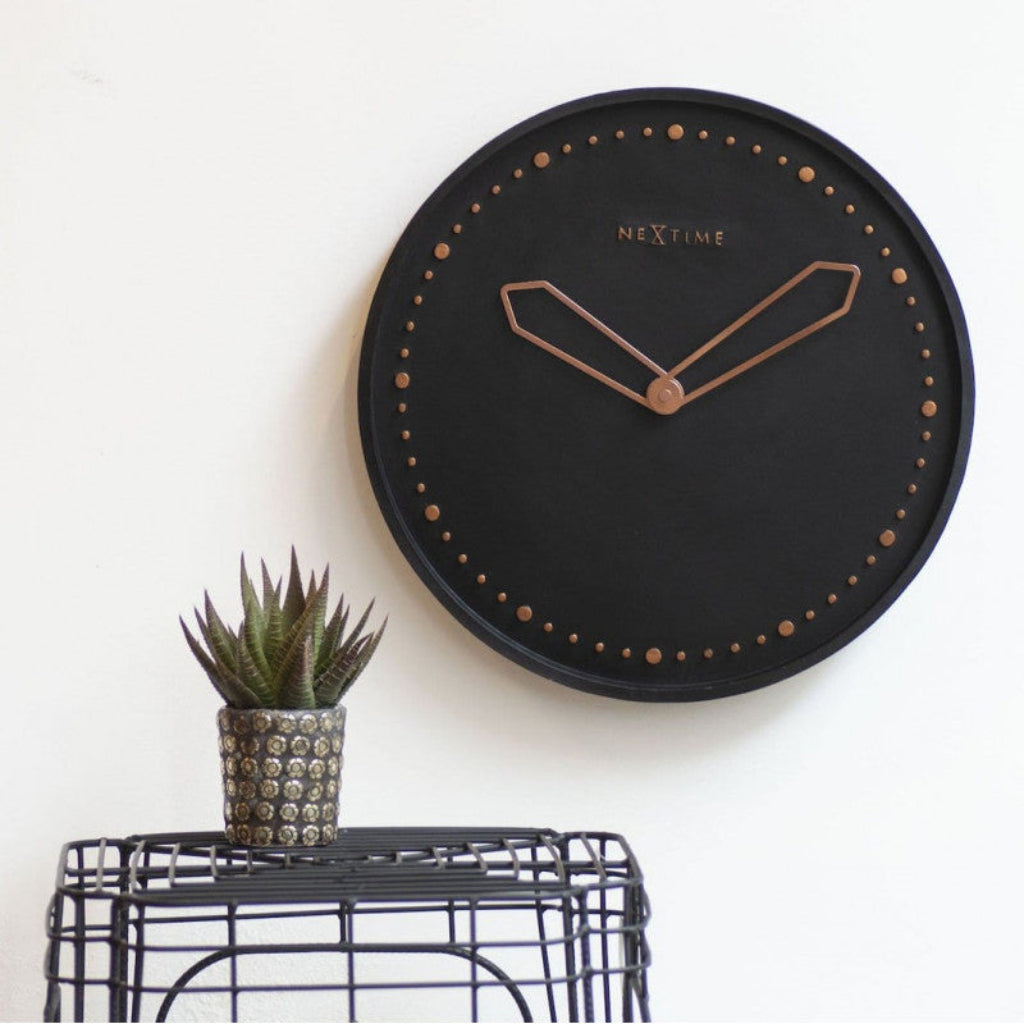 Black wall clock with rose gold hands on a white wall, next to a small potted plant.