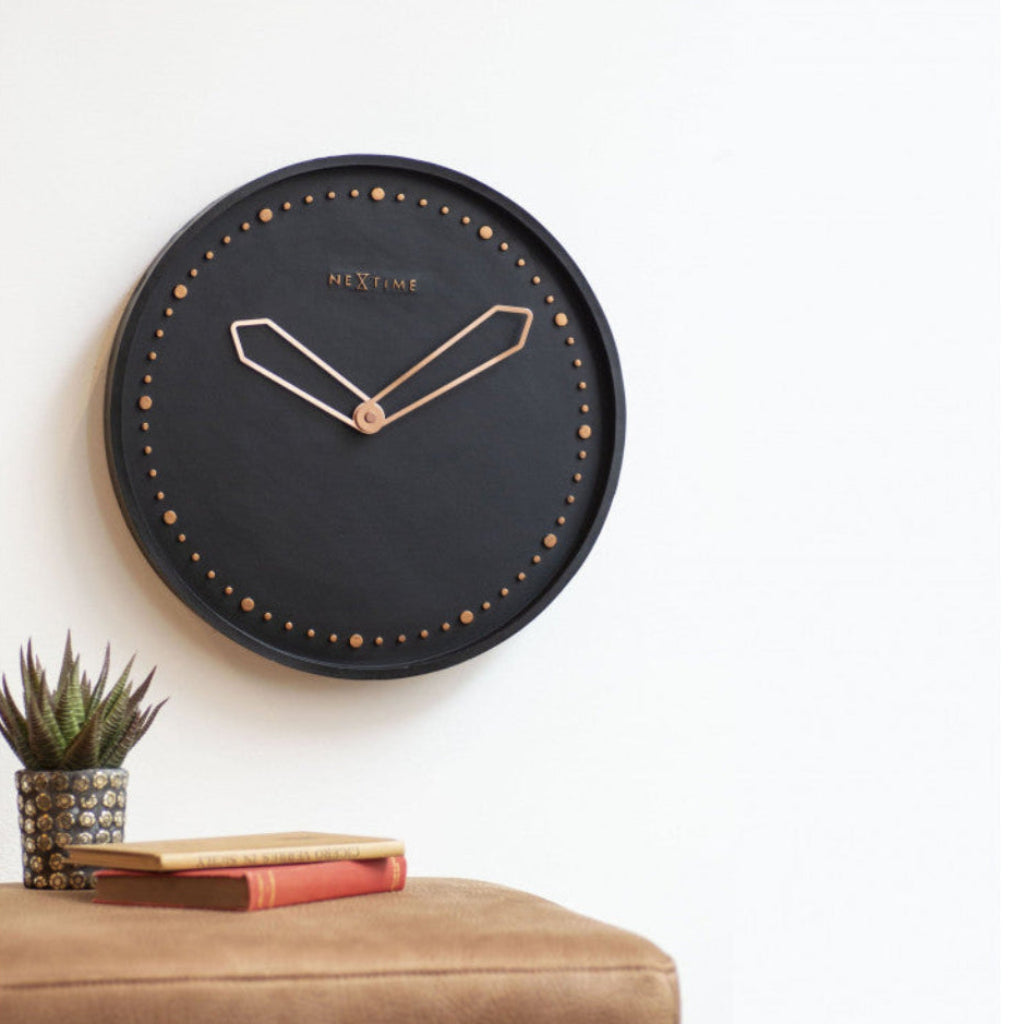 Black wall clock with rose gold accents on a white wall above a brown surface with a plant and book.