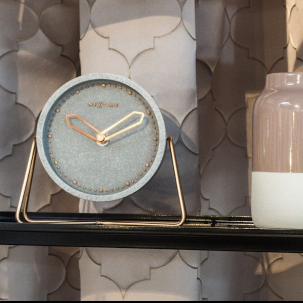 Round clock with gray textured face and gold accents on a stand against a decorative wall.