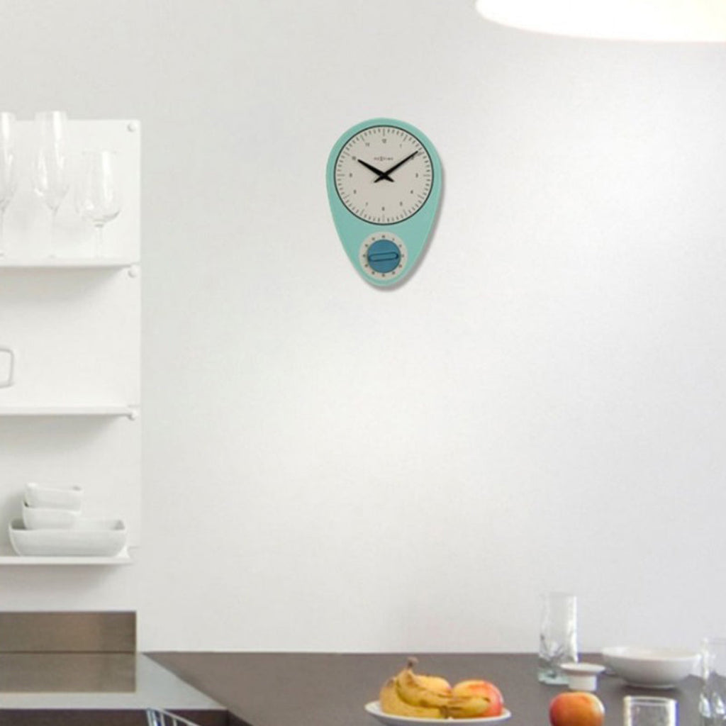 Teal wall clock on a white wall with a kitchen counter and fruit in the foreground.
