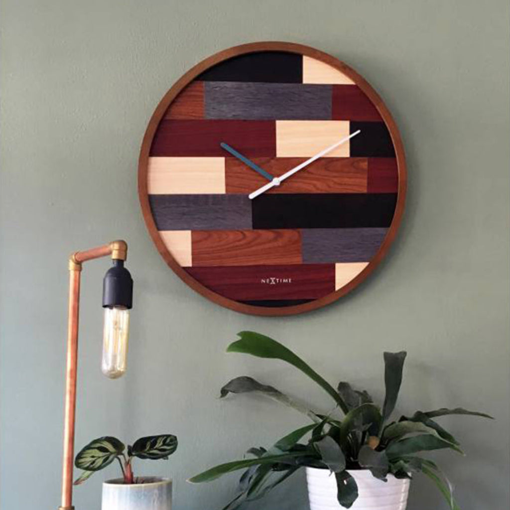 Wooden wall clock with geometric design on a green wall, with a lamp and plants in the foreground.