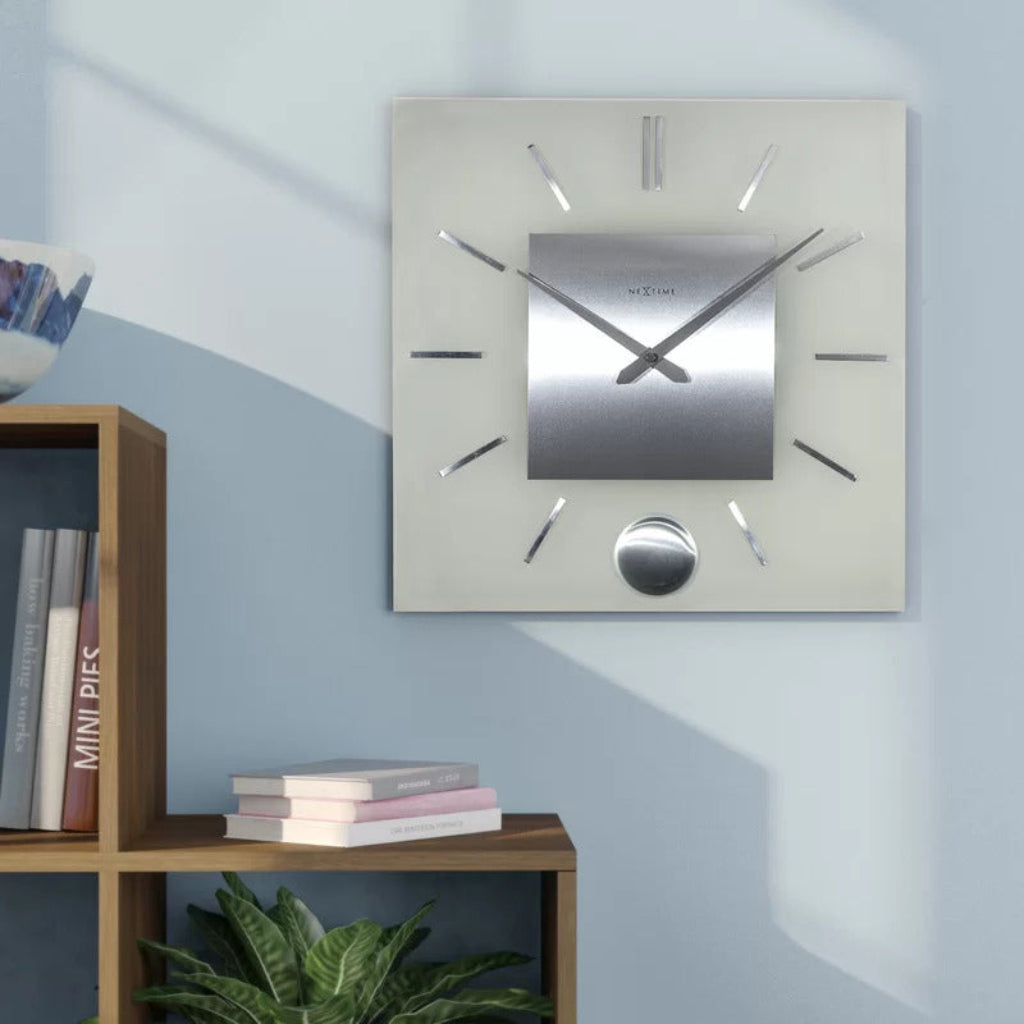 Silver square wall clock on a light blue wall with a wooden shelf and books below.