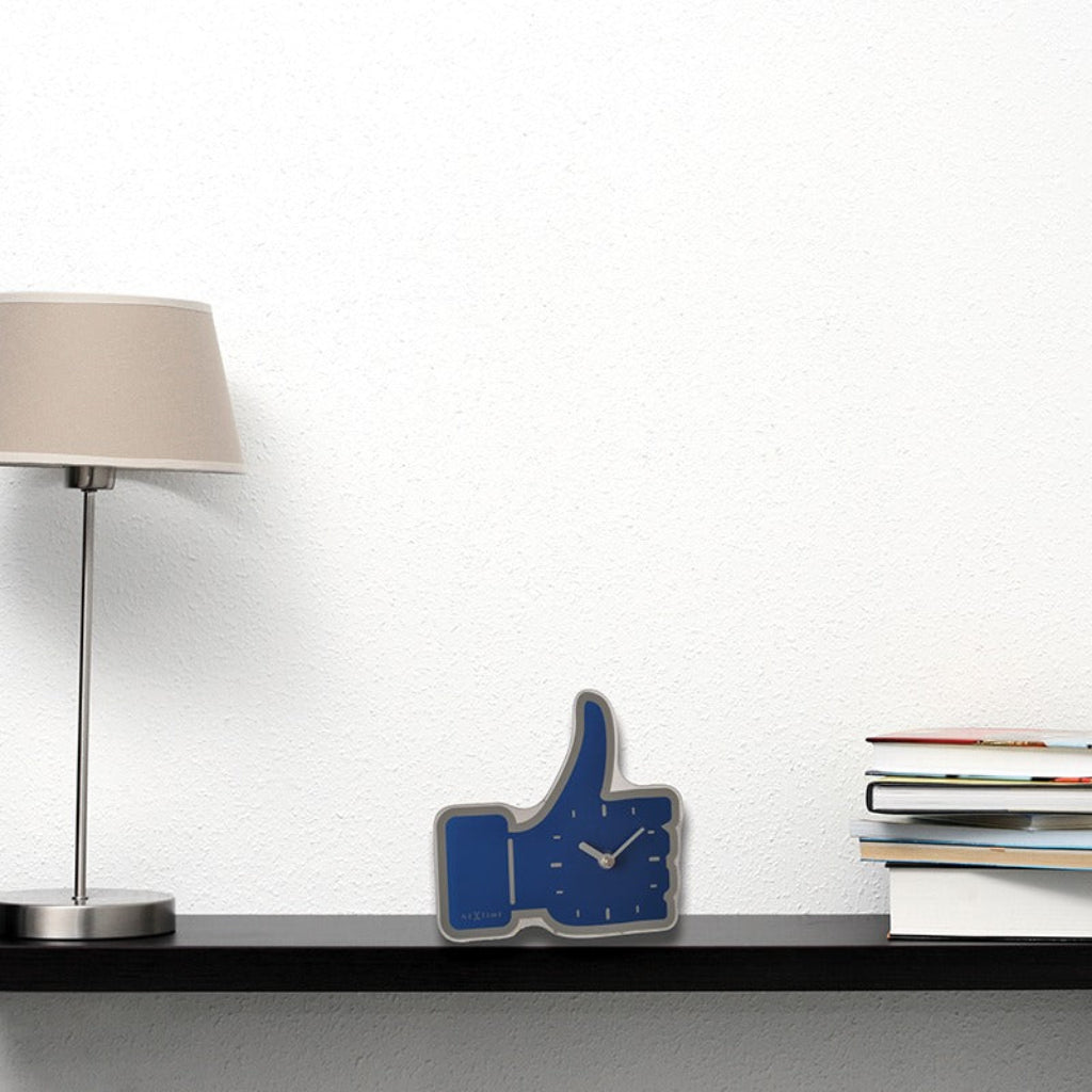 Blue 'like' sign clock on a desk with a lamp and books in the background