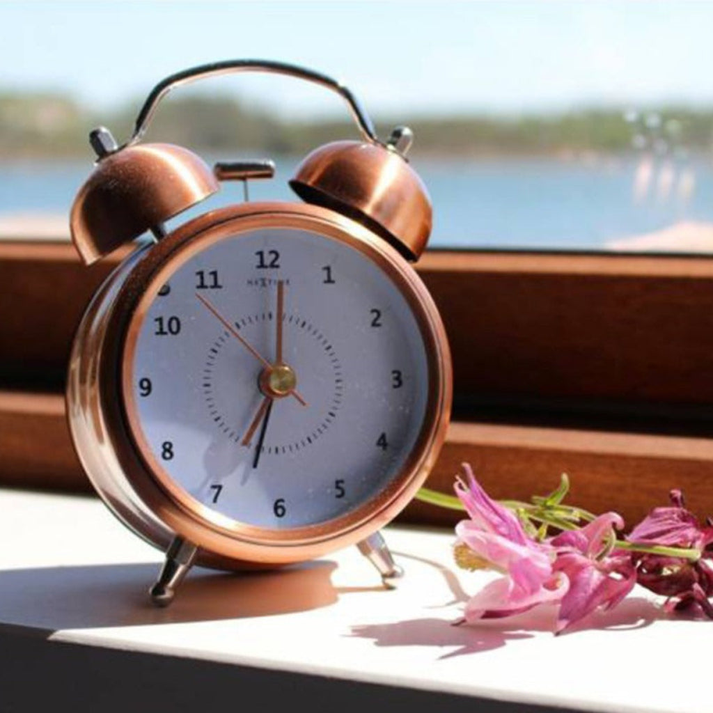 Copper alarm clock on a windowsill with flowers and a blurred outdoor background