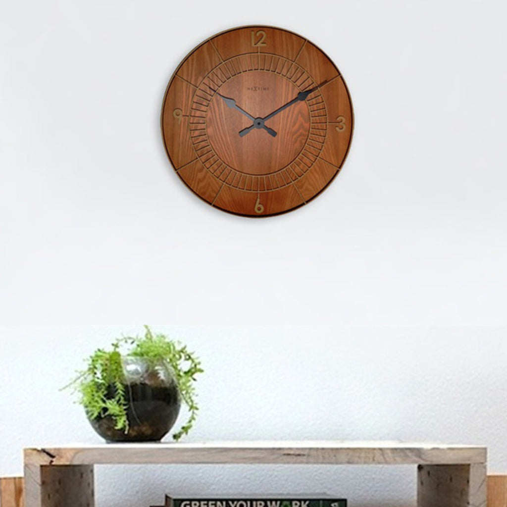Wooden wall clock on a white wall with a small plant on a wooden table below.