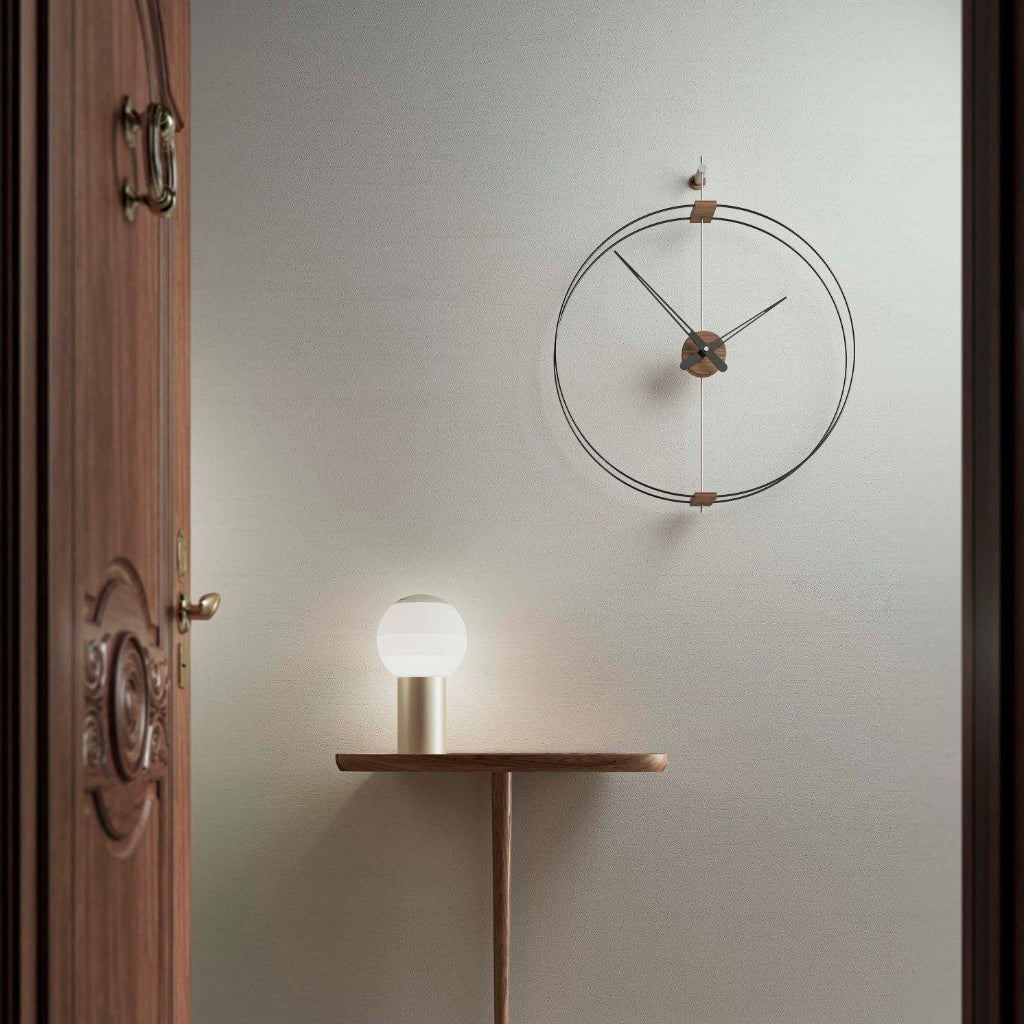 Wall clock on a light gray wall with a small table and lamp in the foreground.
