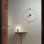 Wall clock on a light gray wall with a small table and lamp in the foreground.