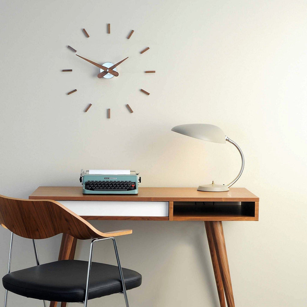 Wooden desk with typewriter, lamp, and chair against a white wall with a clock.