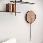Wooden wall clock with pendulum on a white wall above a shelf with books.