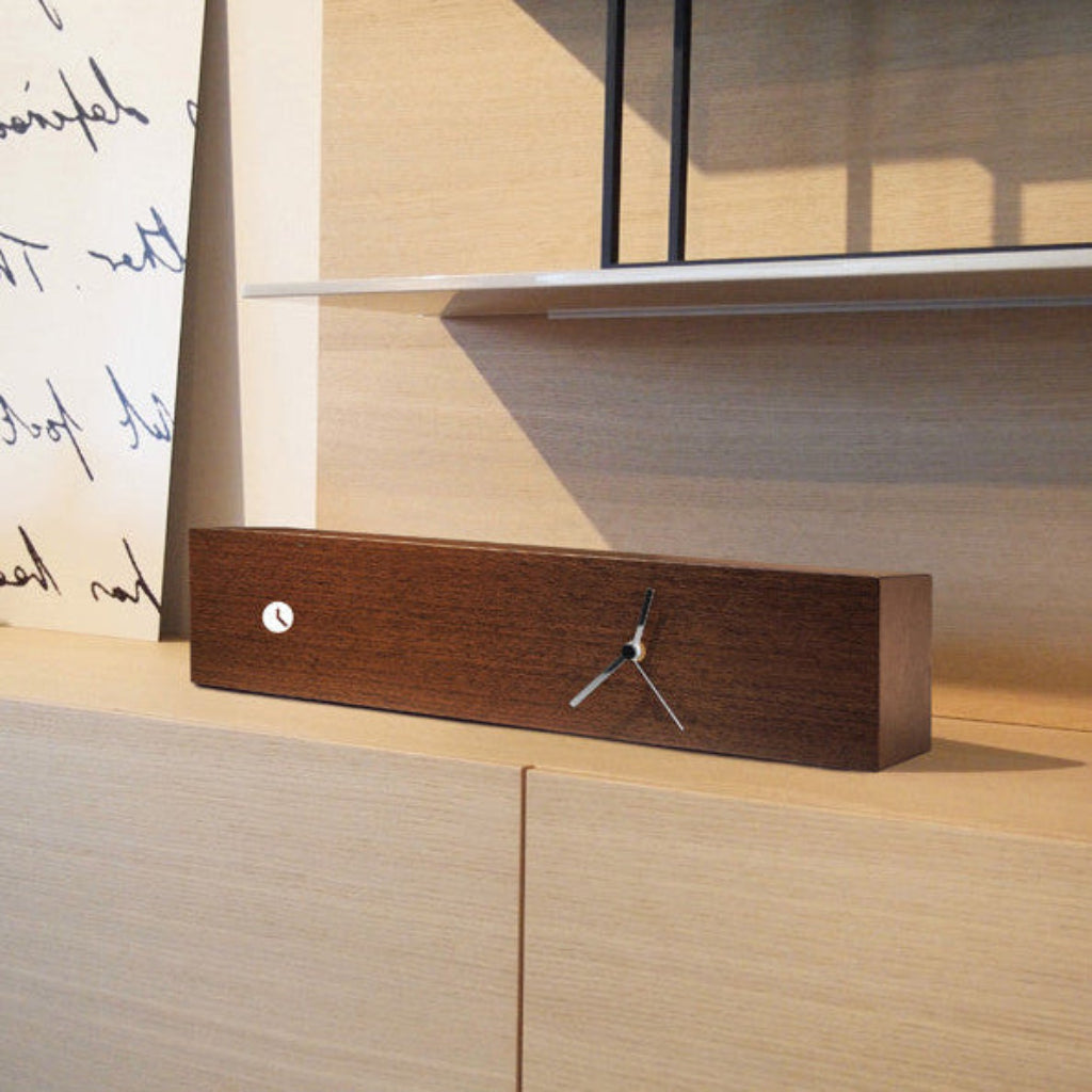 Wooden clock on a wooden shelf with a lamp in the background