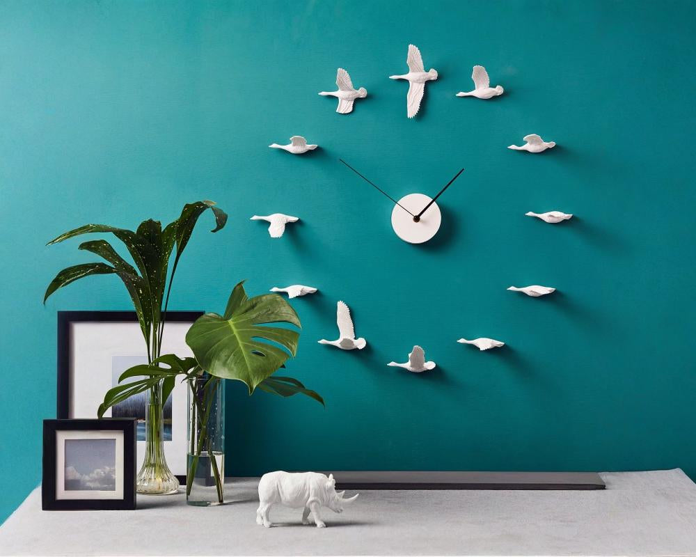 Decorative wall clock with bird designs on a teal wall, with a plant and small figurines in the foreground.
